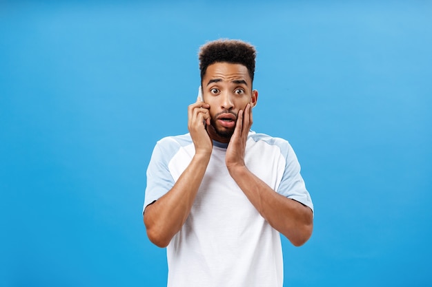 Uomo che prende il telefono sentendo notizie terribili sentirsi scioccato e preoccupato tenendo lo smartphone vicino all'orecchio aprendo la bocca, accigliato e toccando il viso dallo shock preoccupante per la salute dell'amico sul muro blu.