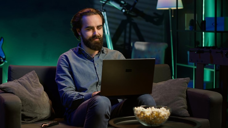 Foto gratuito uomo che scrive sul portatile seduto sul divano e che guarda la tv