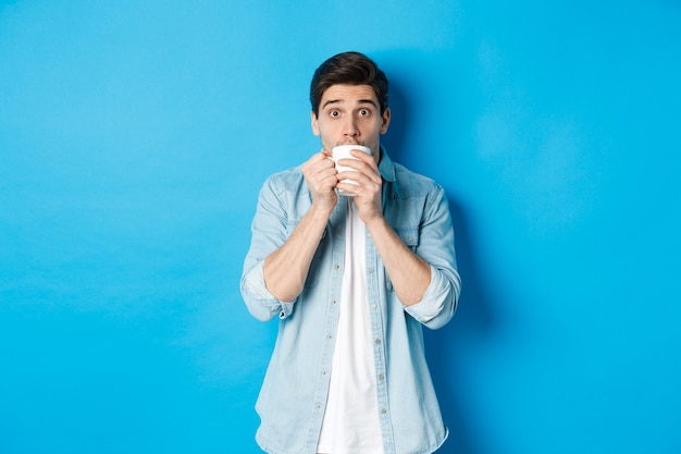 Uomo che sembra eccitato e sorseggia tè o caffè dalla tazza bianca, in piedi su sfondo blu