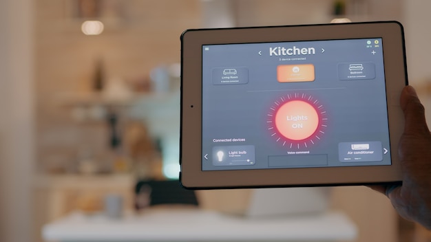 Uomo che tiene in mano un tablet con applicazione di controllo dell'illuminazione che accende le luci seduto in cucina...