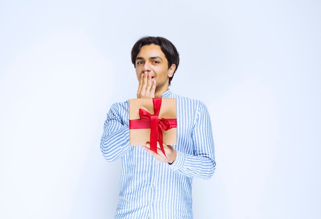 Uomo che tiene una confezione regalo di cartone con nastro rosso e che indica la bocca come sorpreso. Foto di alta qualità