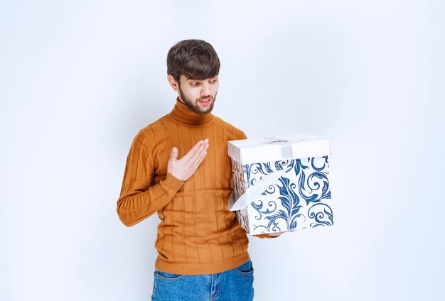 Uomo che tiene una scatola regalo bianca con motivi blu e si indica con sorpresa.