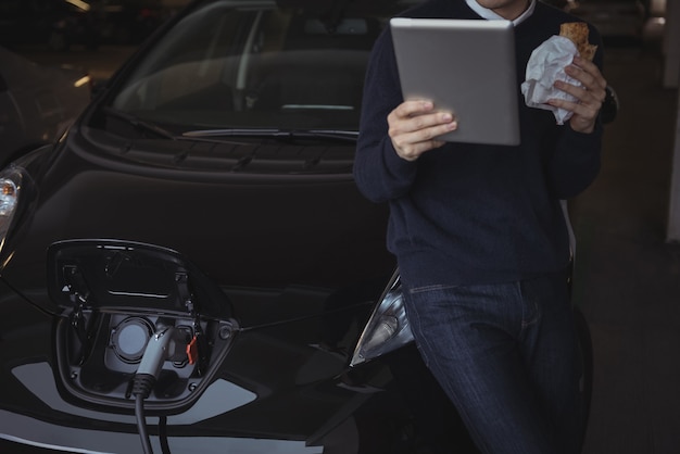 Uomo che utilizza la tavoletta digitale durante la ricarica dell'auto elettrica