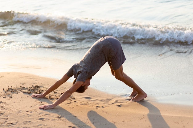 Uomo completo del colpo che pratica yoga sulla spiaggia