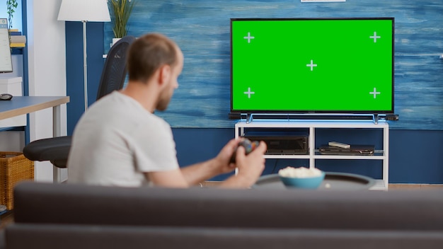 Uomo con controller wireless che gioca con il videogioco per console su una tv a schermo verde mentre è seduto sul divano nel soggiorno moderno. Il giocatore si rilassa sul divano godendosi il gioco online sul display croma key.