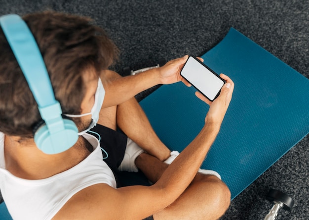 Uomo con cuffie e mascherina medica guardando smartphone in palestra