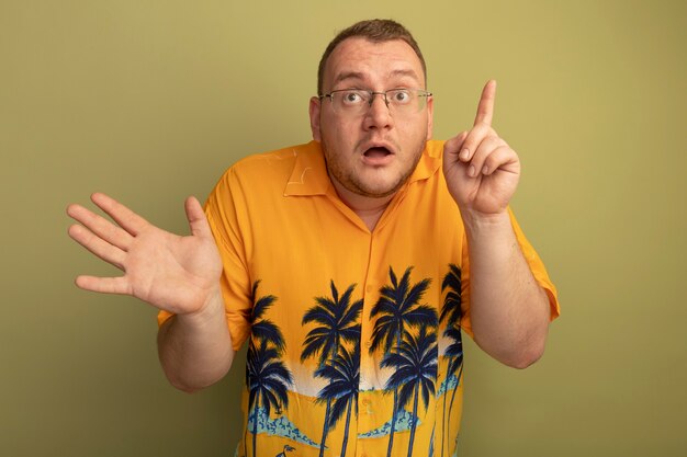 Uomo con gli occhiali che indossa la maglietta arancione che sembra sorpreso che mostra il dito indice in piedi sopra la parete chiara