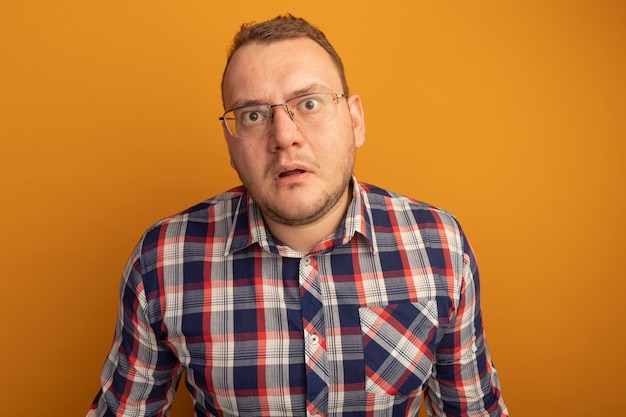 Uomo con gli occhiali e la camicia a quadri che è confuso e preoccupato in piedi sopra il muro arancione