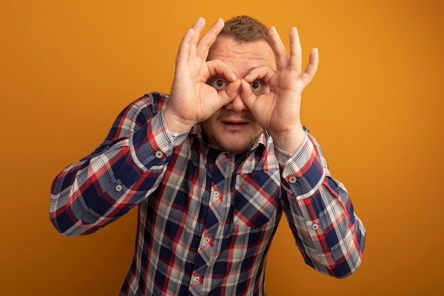 Uomo con gli occhiali e la camicia controllata attraverso le dita che fanno il gesto del binocolo in piedi sopra la parete arancione