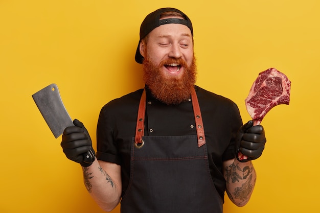 Uomo con la barba allo zenzero in grembiule e guanti che tengono carne e coltello