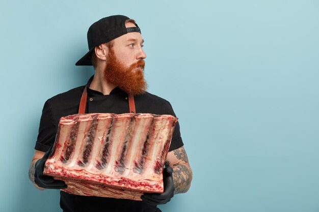 Uomo con la barba allo zenzero in grembiule e guanti che tengono carne