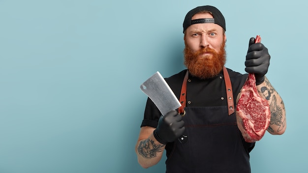 Uomo con la barba allo zenzero in grembiule e guanti che tengono coltello e carne