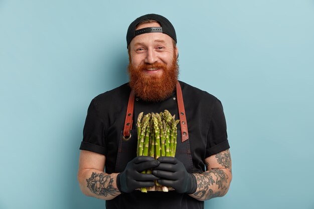 Uomo con la barba allo zenzero in grembiule e guanti che tengono gli asparagi