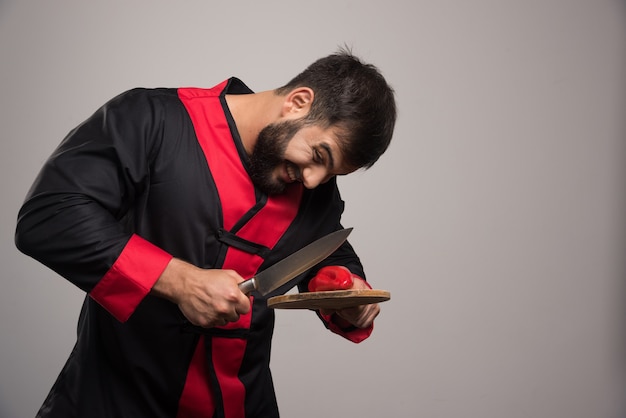 Uomo con la barba che prova a tagliare un peperone rosso sulla tavola di legno.