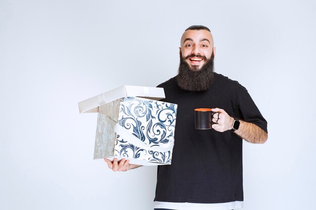 Uomo con la barba che tiene una scatola regalo blu aperta con una tazza di caffè e sembra felice.