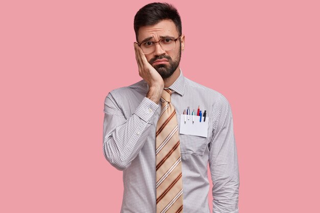 Uomo con la barba lunga abbattuto tiene la mano sulla guancia, indossa abiti formali, ha le penne in tasca, guarda con un'espressione infelice