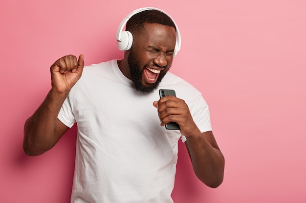 Uomo con la barba lunga nero eccitato canta musica, si muove attivamente, indossa cuffie e maglietta casual, posa su sfondo rosa, tiene la bocca ben aperta