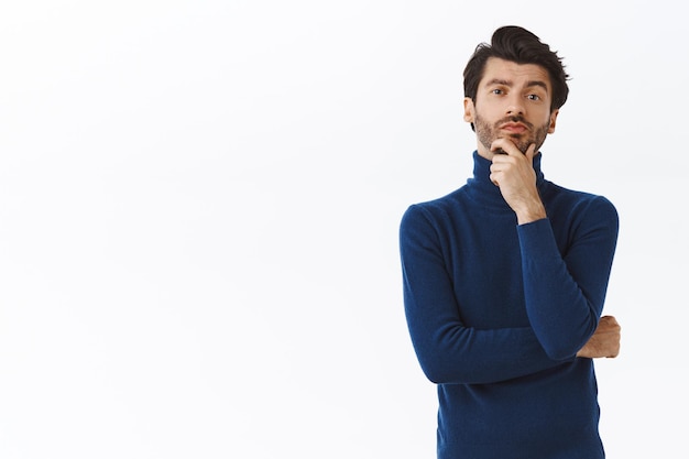 uomo con le setole in maglione alla moda a collo alto, toccando il mento, strofinando la barba e sorridendo mentre giudica qualcosa