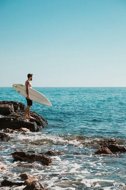 Uomo con tavola da surf in piedi sulla riva sassosa