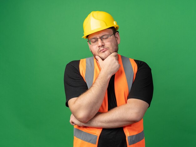 Uomo costruttore in giubbotto da costruzione e casco di sicurezza che guarda con espressione pensosa con la mano sul mento pensando in piedi sopra il verde