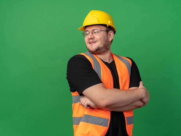 Uomo costruttore in giubbotto da costruzione e casco di sicurezza che guarda da parte con un sorriso sul viso con le braccia incrociate