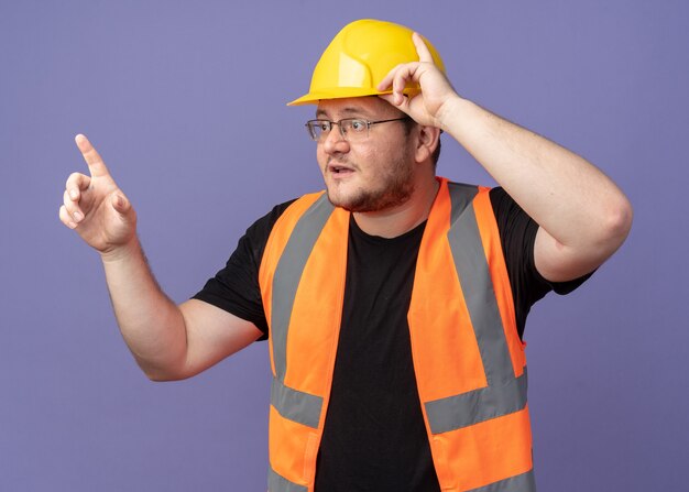 Uomo costruttore in giubbotto da costruzione e casco di sicurezza che guarda da parte preoccupato puntando con il dito indice a qualcosa che sta sopra il blu