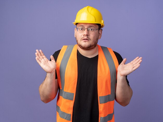 Uomo costruttore in giubbotto da costruzione e casco di sicurezza guardando la telecamera confusa allargando le braccia ai lati in piedi sopra il blu