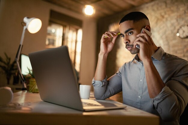 Uomo d'affari afroamericano serio che effettua una telefonata mentre lavora al computer portatile di notte in ufficio