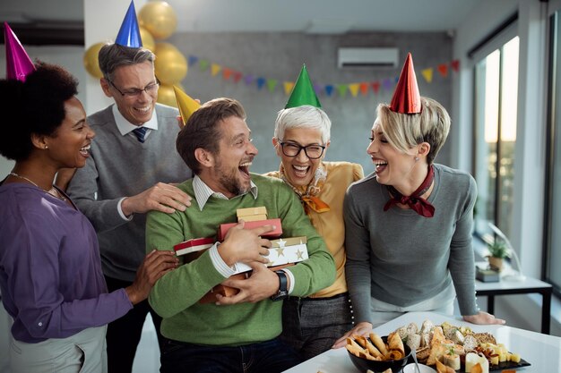 Uomo d'affari allegro che tiene un mazzo di regali di compleanno e si diverte con i colleghi alla festa in ufficio