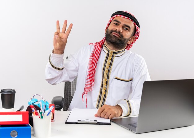 Uomo d'affari arabo in abbigliamento tradizionale seduto al tavolo con computer portatile sorridente mostrando e rivolto verso l'alto con le dita numero tre che lavorano in ufficio