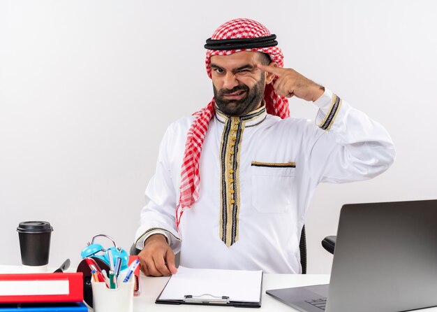 Uomo d'affari arabo in abbigliamento tradizionale seduto al tavolo con il computer portatile che fa l'occhiolino indicando il suo tempio concentrato su un compito che lavora in ufficio