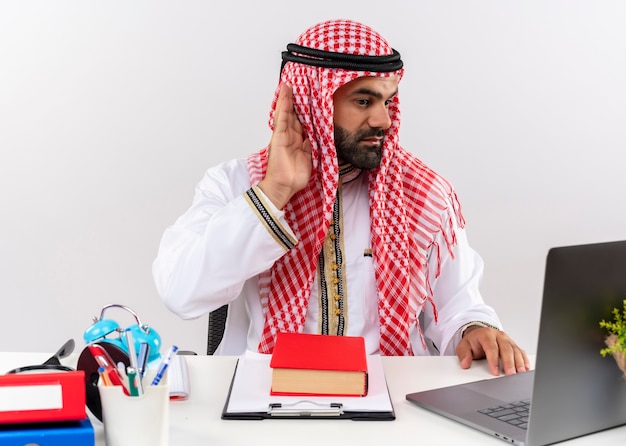 Uomo d'affari arabo nell'usura tradizionale che lavora con il computer portatile che tiene la mano al suo orecchio cercando di ascoltare seduto al tavolo in ufficio