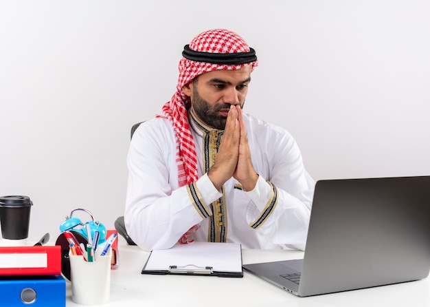 Uomo d'affari arabo nell'usura tradizionale che lavora con il computer portatile che tiene le mani insieme all'espressione di speranza che si siede al tavolo in ufficio