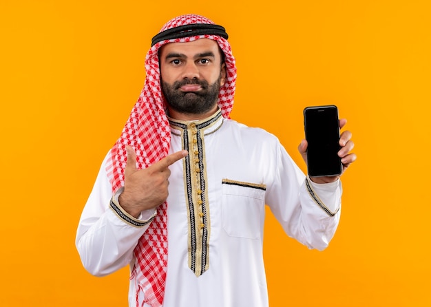 Uomo d'affari arabo nell'usura tradizionale che mostra smartphone che indica con il dito che sembra confuso in piedi sopra la parete arancione