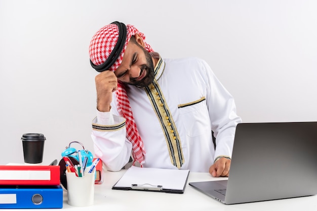 Uomo d'affari arabo nell'usura tradizionale che si siede al tavolo con il computer portatile che sembra stanco e deluso lavorando in ufficio