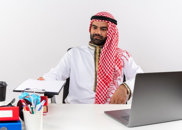Uomo d'affari arabo nell'usura tradizionale che si siede al tavolo con il computer portatile che tiene appunti con pagine vuote che lavorano in ufficio