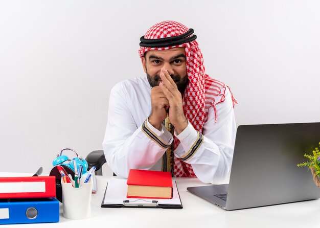 Uomo d'affari arabo nell'usura tradizionale che si siede al tavolo con il computer portatile sorridendo furbescamente felice e soddisfatto di lavorare in ufficio