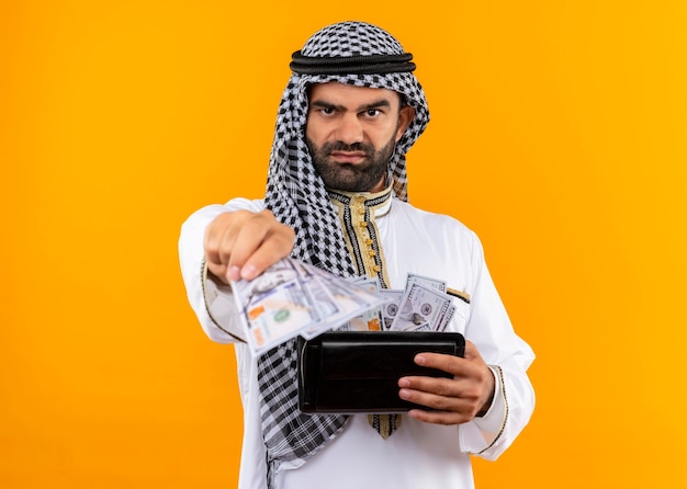 Uomo d'affari arabo nell'usura tradizionale che tiene portafoglio con contanti con la faccia seria che sta sopra la parete arancione
