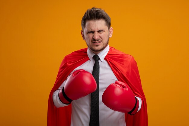 Uomo d'affari arrabbiato super eroe in mantello rosso e guantoni da boxe che guarda l'obbiettivo con espressione aggressiva pronta a combattere in piedi su sfondo arancione