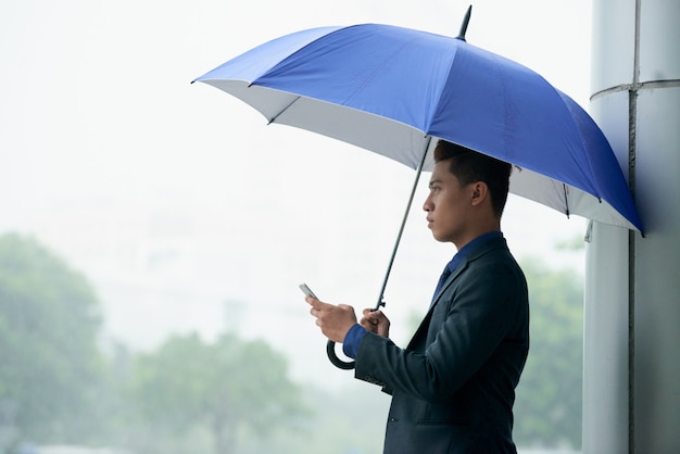 Uomo d'affari asiatico che sta in via con l'ombrello durante la pioggia e che per mezzo dello smartphone