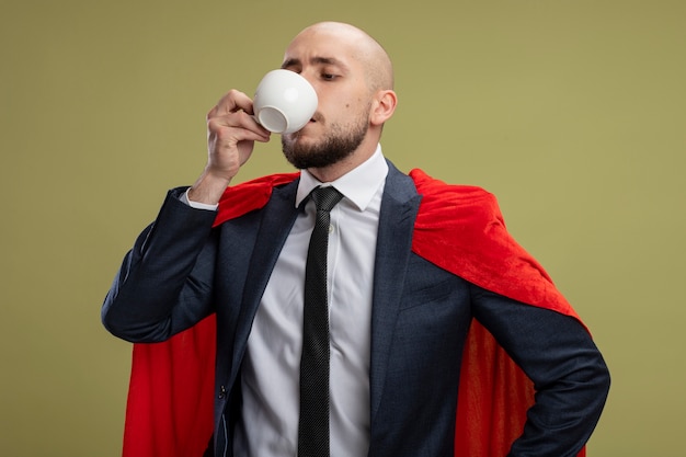 Uomo d'affari barbuto super eroe in mantello rosso che beve caffè guardando fiducioso in piedi sopra la parete verde chiaro