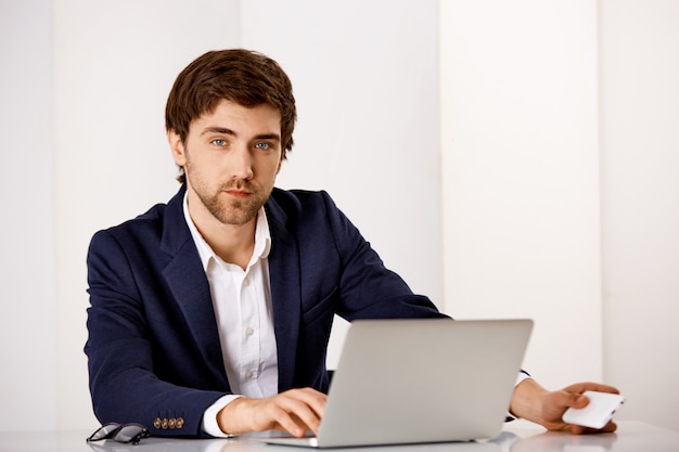 Uomo d'affari bello dall'aspetto serio in tuta, sedersi alla scrivania, lavorare sul rapporto con il computer portatile, attendere la telefonata