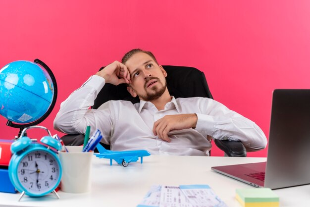 Uomo d'affari bello in camicia bianca che osserva in su con sguardo sognante che si siede al tavolo in ufficio su sfondo rosa