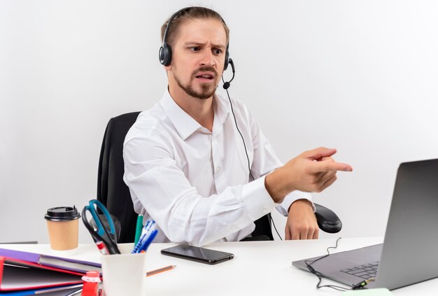 Uomo d'affari bello in camicia bianca e cuffie con un microfono che lavora al computer portatile che sembra confuso seduto al tavolo in ufficio su sfondo bianco