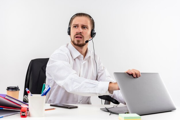 Uomo d'affari bello in camicia bianca e cuffie con un microfono che lavora su un computer portatile che osserva da parte con la faccia seria che si siede al tavolo in offise sopra priorità bassa bianca