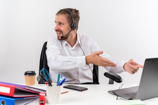 Uomo d'affari bello in camicia bianca e cuffie con un microfono che lavora su un computer portatile che osserva da parte confuso seduto al tavolo in ufficio su sfondo bianco