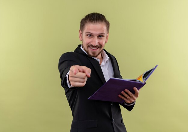 Uomo d'affari bello in taccuino della tenuta del vestito nero che guarda l'obbiettivo che indica con il dito sorridente che sta sopra fondo oliva