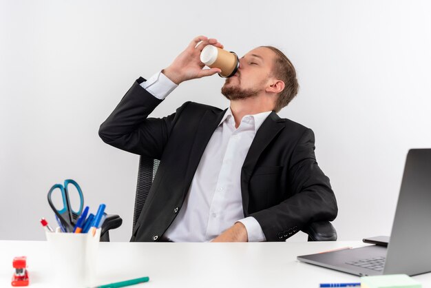 Uomo d'affari bello in vestito che beve caffè che si siede al tavolo in carica su sfondo bianco