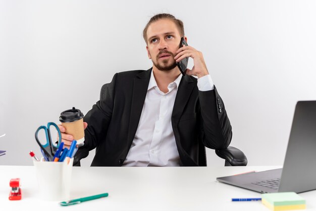 Uomo d'affari bello in vestito che lavora al computer portatile che parla sul telefono cellulare che osserva da parte con la faccia seria che si siede al tavolo in ufficio sopra priorità bassa bianca