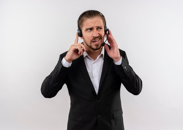 Uomo d'affari bello in vestito e cuffie con un microfono che ascolta un cliente che sembra confuso in piedi su sfondo bianco